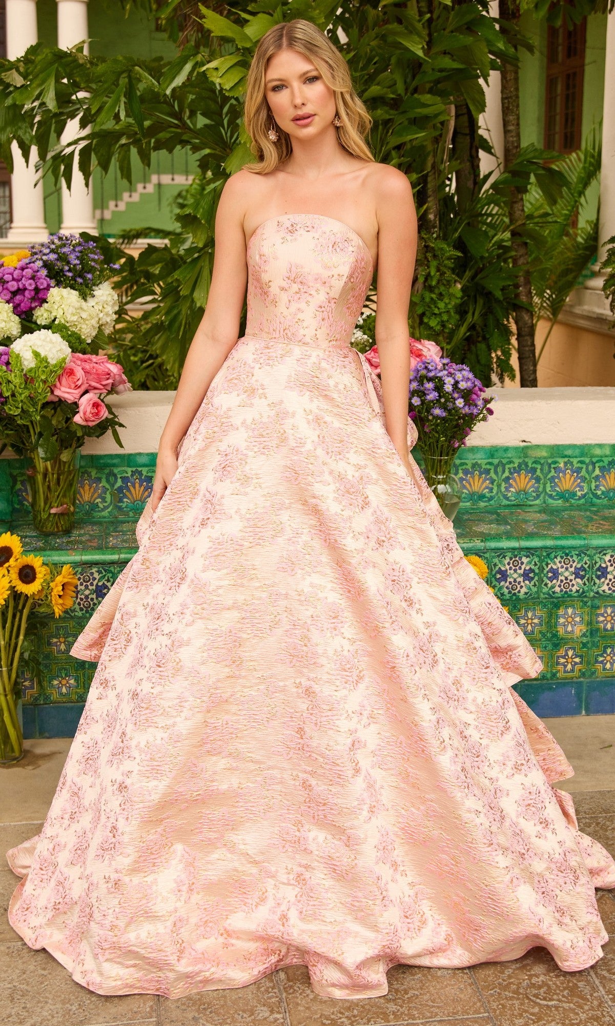A woman stands outdoors before green foliage and flowers, wearing the Ava Presley 26412 Ruffled Strapless Prom Gown in blush, an A-line evening dress with subtle floral design.